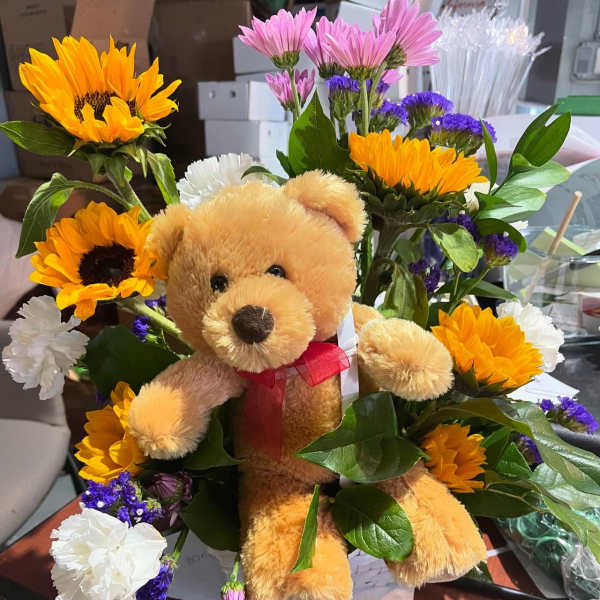 Stuffed teddy bear nestled among yellow sunflowers, white carnations, and pink and purple daisies.