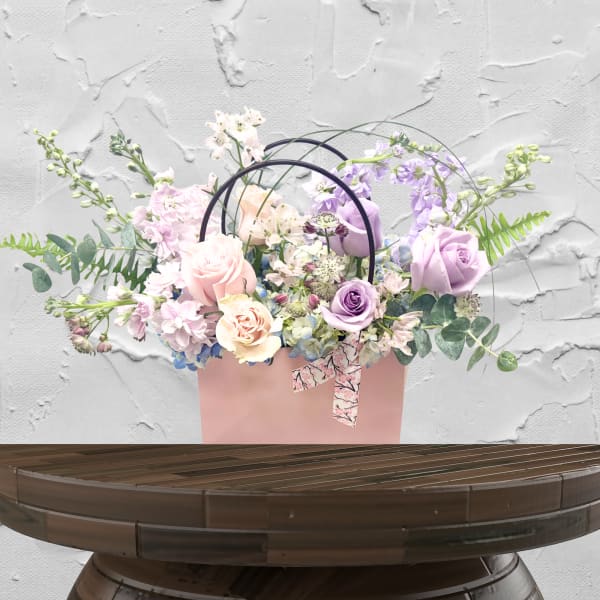 Pastel roses and lavender flowers arranged in a pink gift bag with ribbon handles.