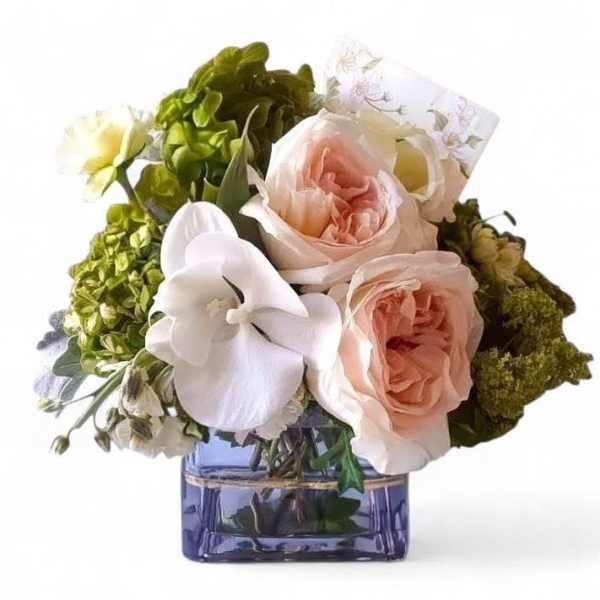 Low arrangement of peach roses and white orchid in a square purple glass vase with a card