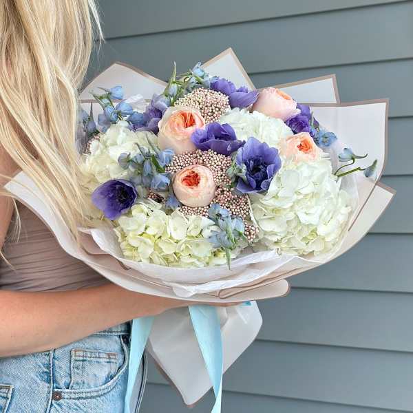 Handheld bouquet of white, peach, and purple flowers wrapped in paper
