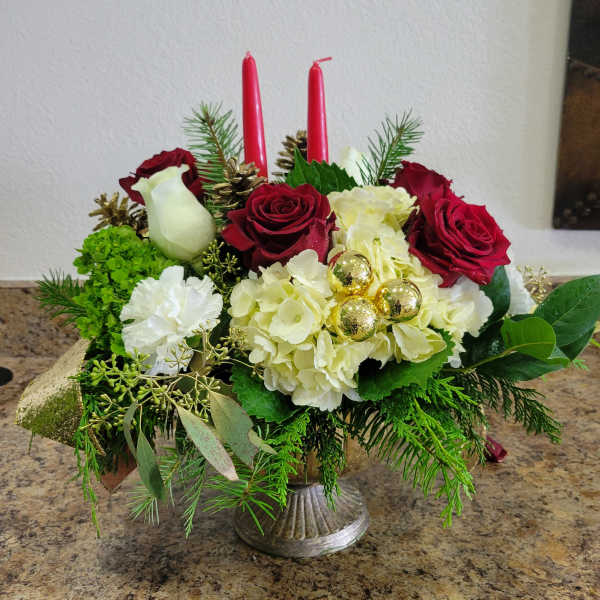 Holiday centerpiece with red roses, white hydrangeas, evergreens, and two red taper candles in a silver pedestal bowl