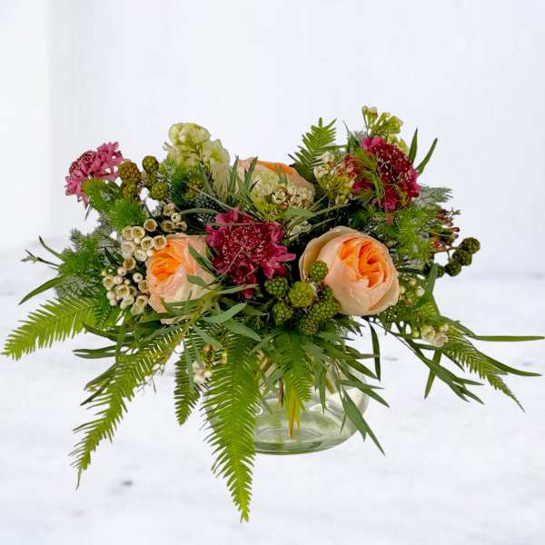 Peach roses and burgundy flowers in a glass vase with fern foliage