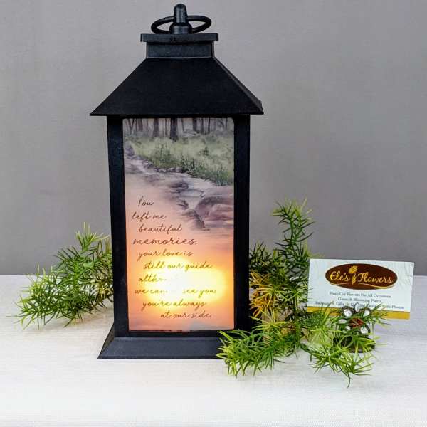 Black memorial lantern with glowing light and verse, accented by small green sprigs.