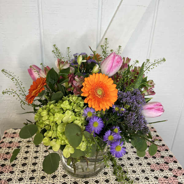 Mixed bouquet with orange gerbera daisies, pink tulips, and purple flowers in a glass vase