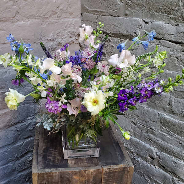 Mixed bouquet of blue, purple, and white flowers in a clear square vase