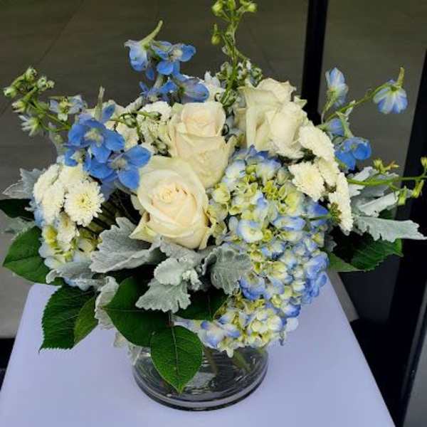 Low round arrangement of cream roses, blue delphinium, hydrangeas, and white mums in a clear glass vase