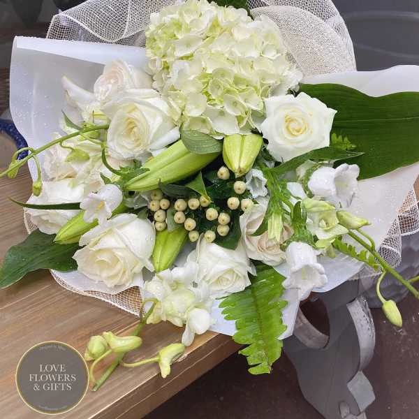 Hand-tied bouquet of white roses, lilies, orchids, and hydrangea wrapped in white mesh and paper