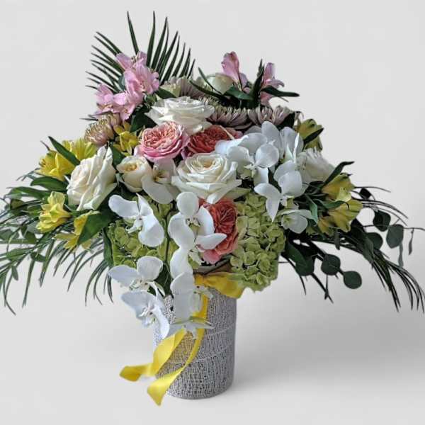 Tall arrangement of white orchids, roses, and mixed blooms in a textured vase with yellow ribbon.