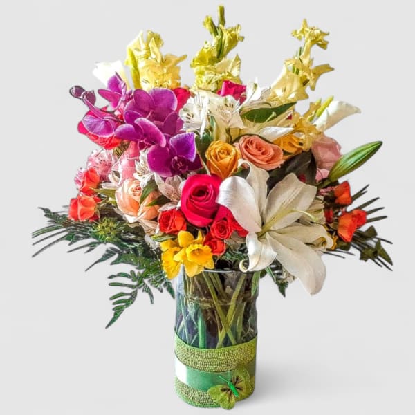 Tall mixed bouquet of bright roses, lilies, orchids, and other blooms in a glass vase with green ribbon.