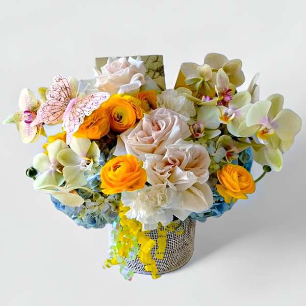 Mixed arrangement of yellow ranunculus, blush roses, orchids, and blue hydrangeas in a metal vase with ribbon