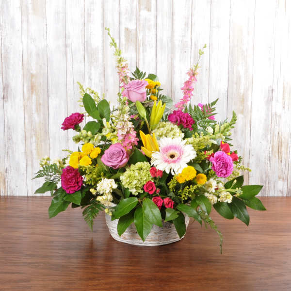 Colorful mixed flower basket with pink roses, yellow lilies, and a white gerbera on a wooden table.