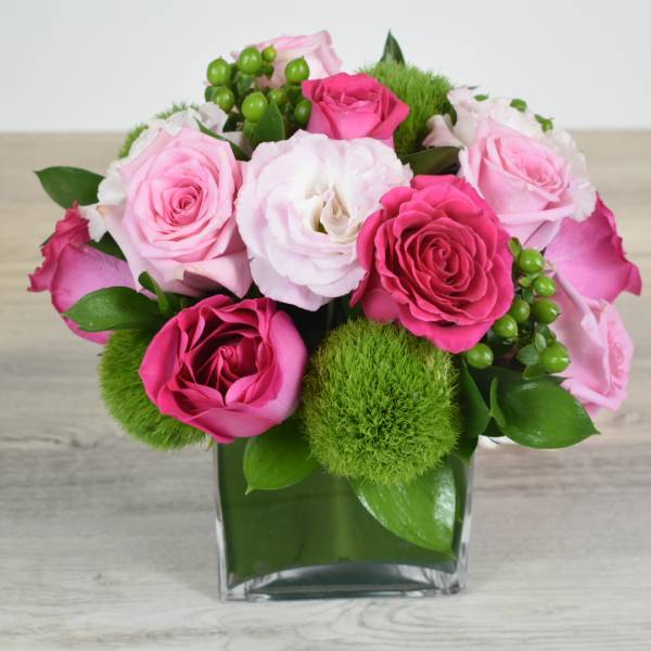 Compact arrangement of pink roses, white lisianthus, and green dianthus in a clear glass cube vase
