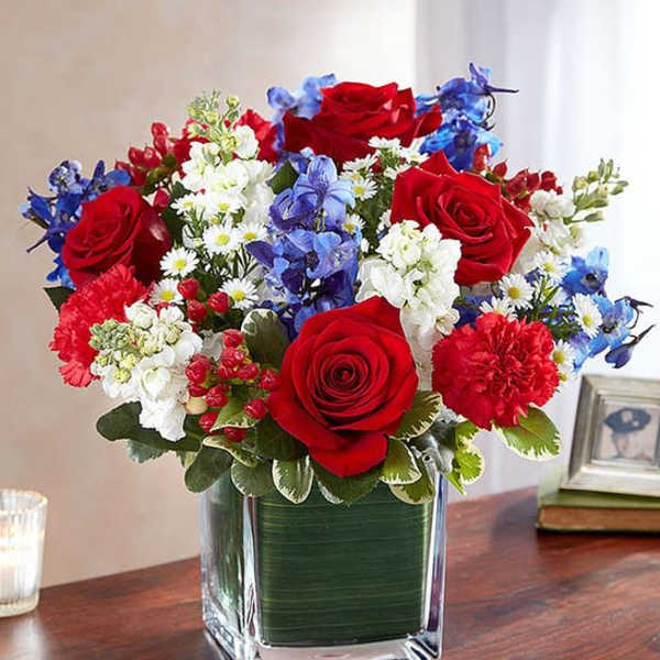 Red roses, blue flowers, and white daisies arranged in a square glass vase