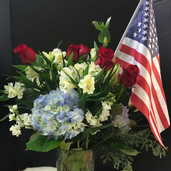 Red roses and white lilies with blue hydrangea in a glass vase beside an American flag