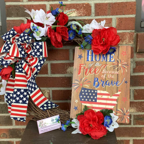 Patriotic wreath with red, white, and blue flowers and a flag ribbon beside a wooden sign