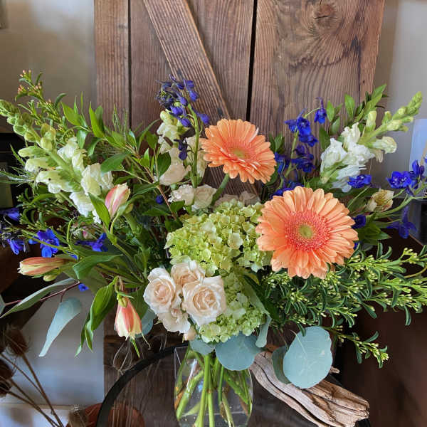 Peach gerbera daisies, cream roses, green hydrangea, and blue delphinium in a clear glass vase