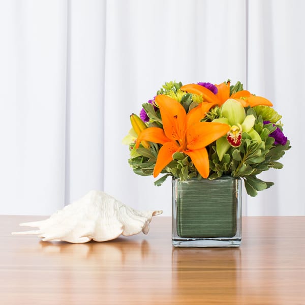 Low arrangement of orange lilies, green orchids, and purple blooms in a glass cube vase beside a white seashell.