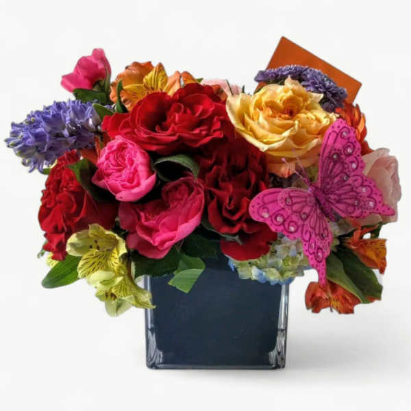 Bright mixed bouquet of roses and other blooms in a square glass vase with a pink butterfly pick