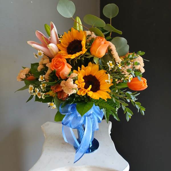 Orange and yellow bouquet with sunflowers, roses, lilies, and a blue ribbon in a vase