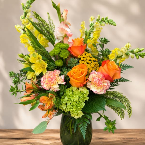 Mixed bouquet of orange roses, yellow snapdragons, and carnations in a green glass vase