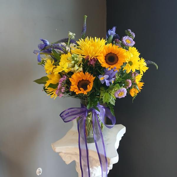 Bouquet of sunflowers, yellow daisies, and purple flowers in a glass vase with a purple ribbon