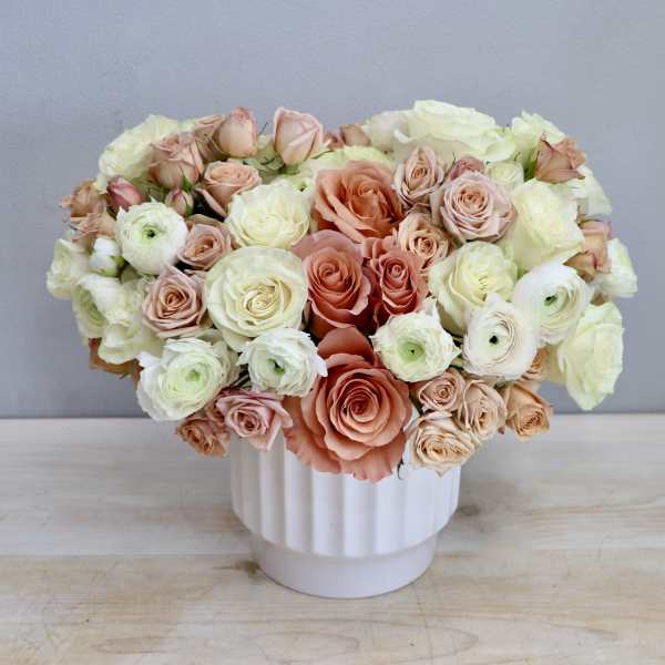 Low arrangement of peach and cream roses with ranunculus in a white ceramic vase