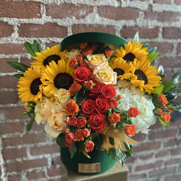 Round green hatbox arrangement with yellow sunflowers, orange and red roses, and white hydrangeas.