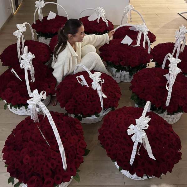 A woman sits among large white baskets overflowing with deep red roses and white bows.