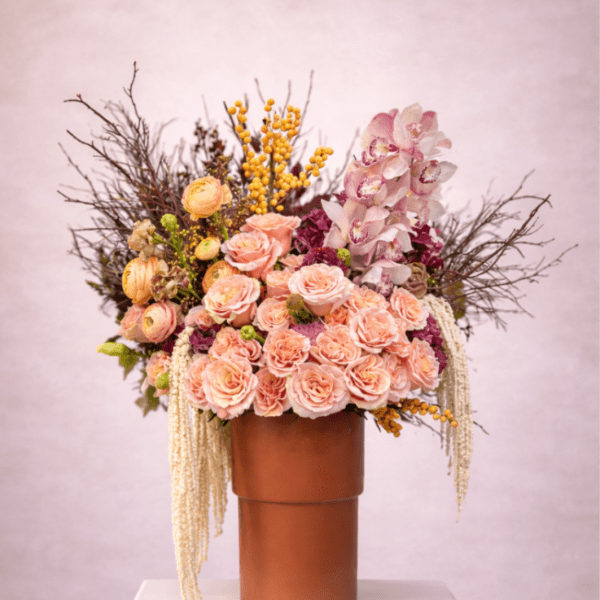 Tall arrangement of peach roses, ranunculus, orchids, and yellow berries in a brown vase on a white pedestal.