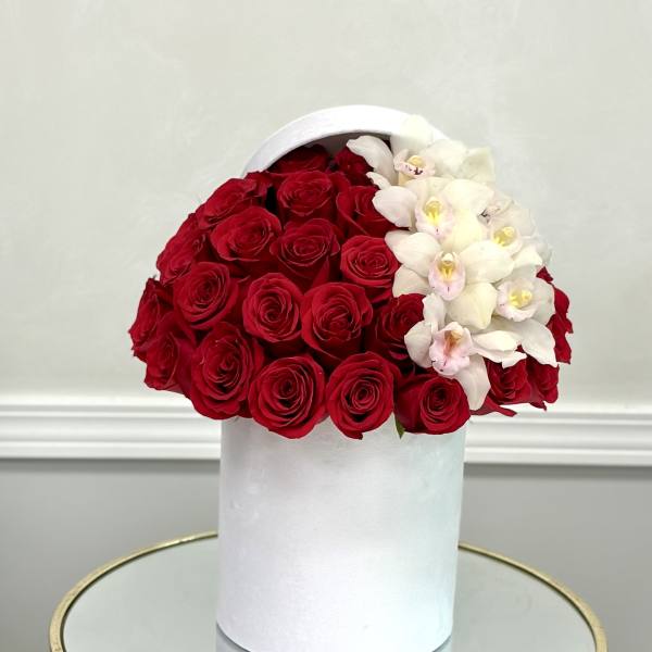 Red roses and white orchids arranged in a white hatbox-style cylinder container on a mirrored table