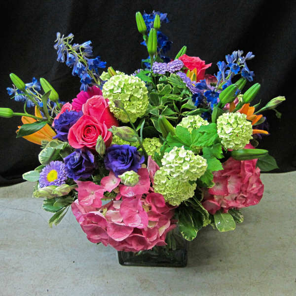 Colorful arrangement of pink hydrangeas, roses, green blooms and mixed purple and blue flowers in a glass cube vase