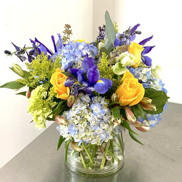 Bouquet of yellow roses, blue hydrangeas, and purple flowers in a glass vase
