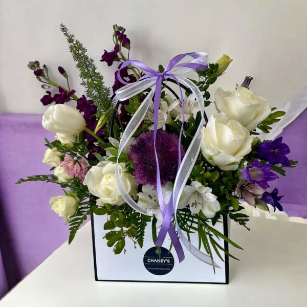 Purple and white floral arrangement in a gift bag with ribbon