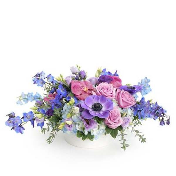 Low arrangement of purple anemones, lavender roses, and blue flowers in a white bowl.
