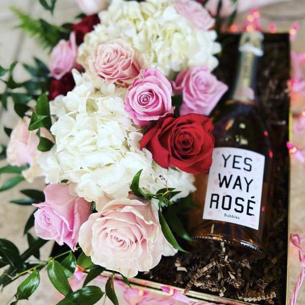 Pink and white roses with hydrangeas beside a bottle of rosé in a gift box