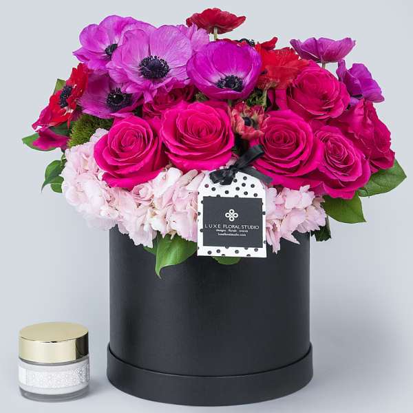 Pink and purple flower arrangement in a black hatbox with a small candle jar beside it.