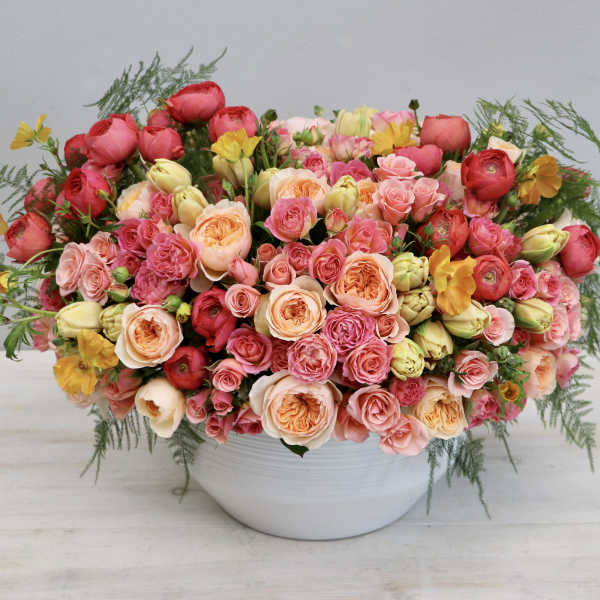 Low white bowl filled with dense pink, peach, and yellow roses and buds in a rounded arrangement.