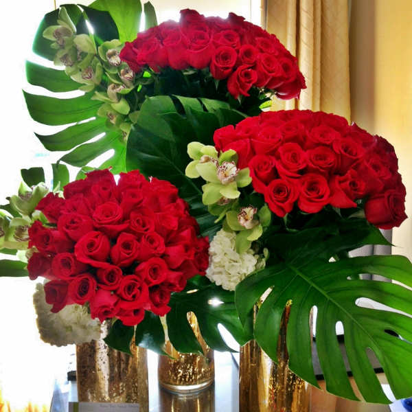 Red rose arrangements with orchids and large tropical leaves in gold vases