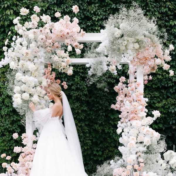 Bride in a white gown beside a floral wedding arch with white and blush flowers