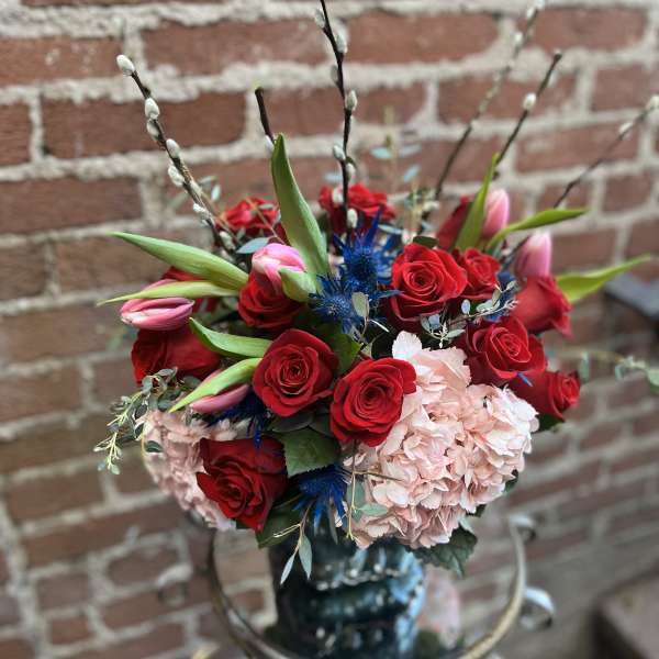Tall arrangement of red roses, pink tulips, and pink hydrangea with blue accent flowers in a dark vase.