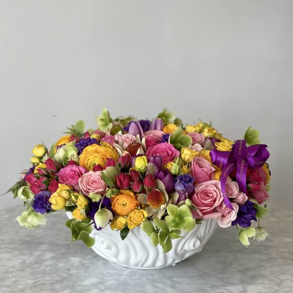 Mixed bouquet of pink, yellow, purple, and green flowers in a white bowl vase