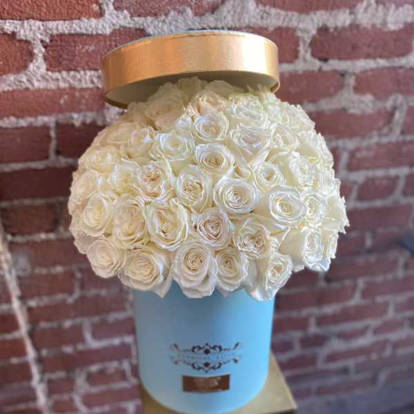Round box of cream roses in a light blue hatbox with a gold lid against a brick wall.