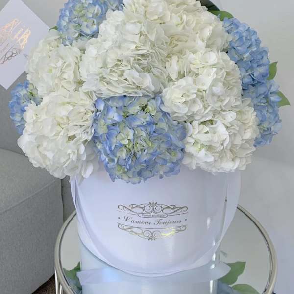 Round box of white and light blue hydrangeas with a card on a mirrored table