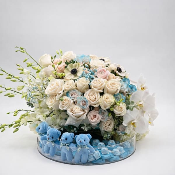 Bouquet of white and pastel roses with orchids in a glass bowl, with blue teddy bears below.