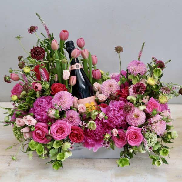Low pink floral arrangement with roses and tulips surrounding a bottle of champagne in a white container.