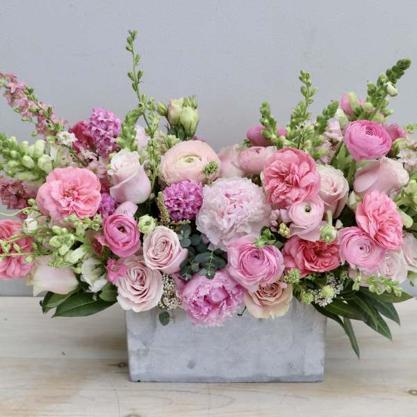 Low rectangular centerpiece of pink roses, ranunculus, peonies and snapdragons in a concrete container.