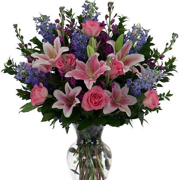 Pink lilies and roses with blue flowers in a clear glass vase