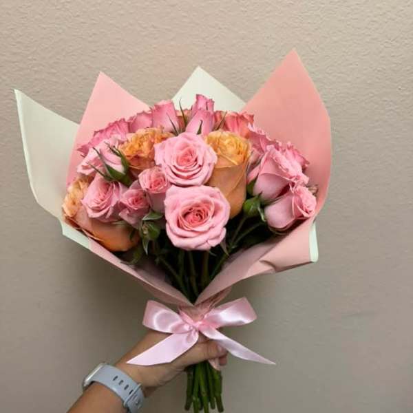 Handheld bouquet of pink and peach roses wrapped in pastel paper with a pink ribbon