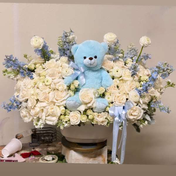 Blue teddy bear in a white rose and blue flower arrangement