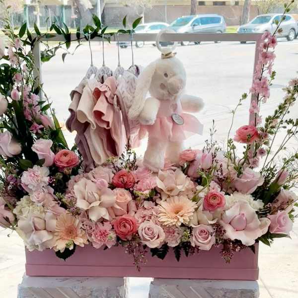 Pink floral display with a teddy bear and baby clothes in a box